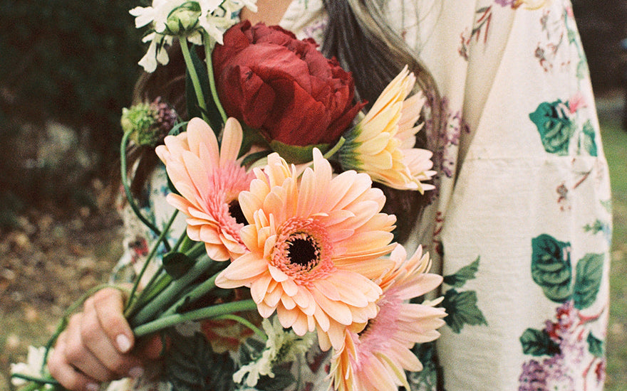 Model holding flowers wearing a Metta Melbourne 'Paloma Dress in Bouquet Print' floral dress. 